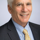 Jared Bernstein stands smiling in front of a grey background wearing a dark grey jacket, yellow striped tie, and light blue shirt.